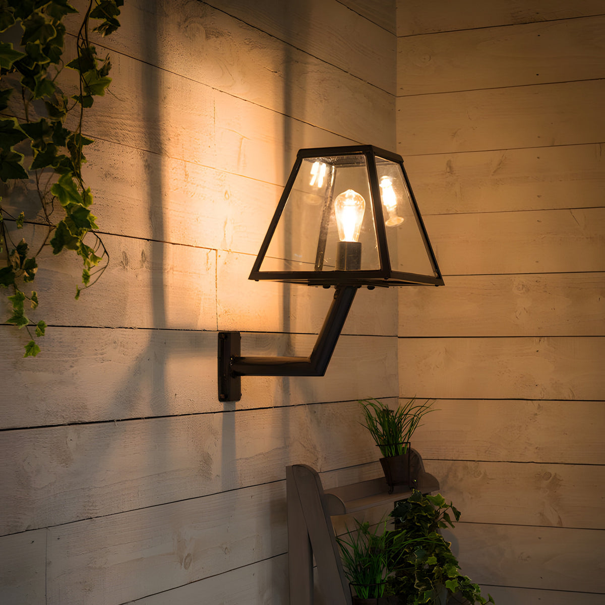 Small contemporary black outdoor wall lantern on an angled bracket, tapered glass shade with visible bulb, mounted on wooden panelled wall near plants.