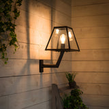 Small contemporary black outdoor wall lantern on an angled bracket, tapered glass shade with visible bulb, mounted on wooden panelled wall near plants.