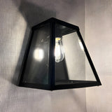 Contemporary Galvanised Steel Bulkhead Wall Light in black, tapered glass panels with visible bulb, mounted against a light grey textured wall.