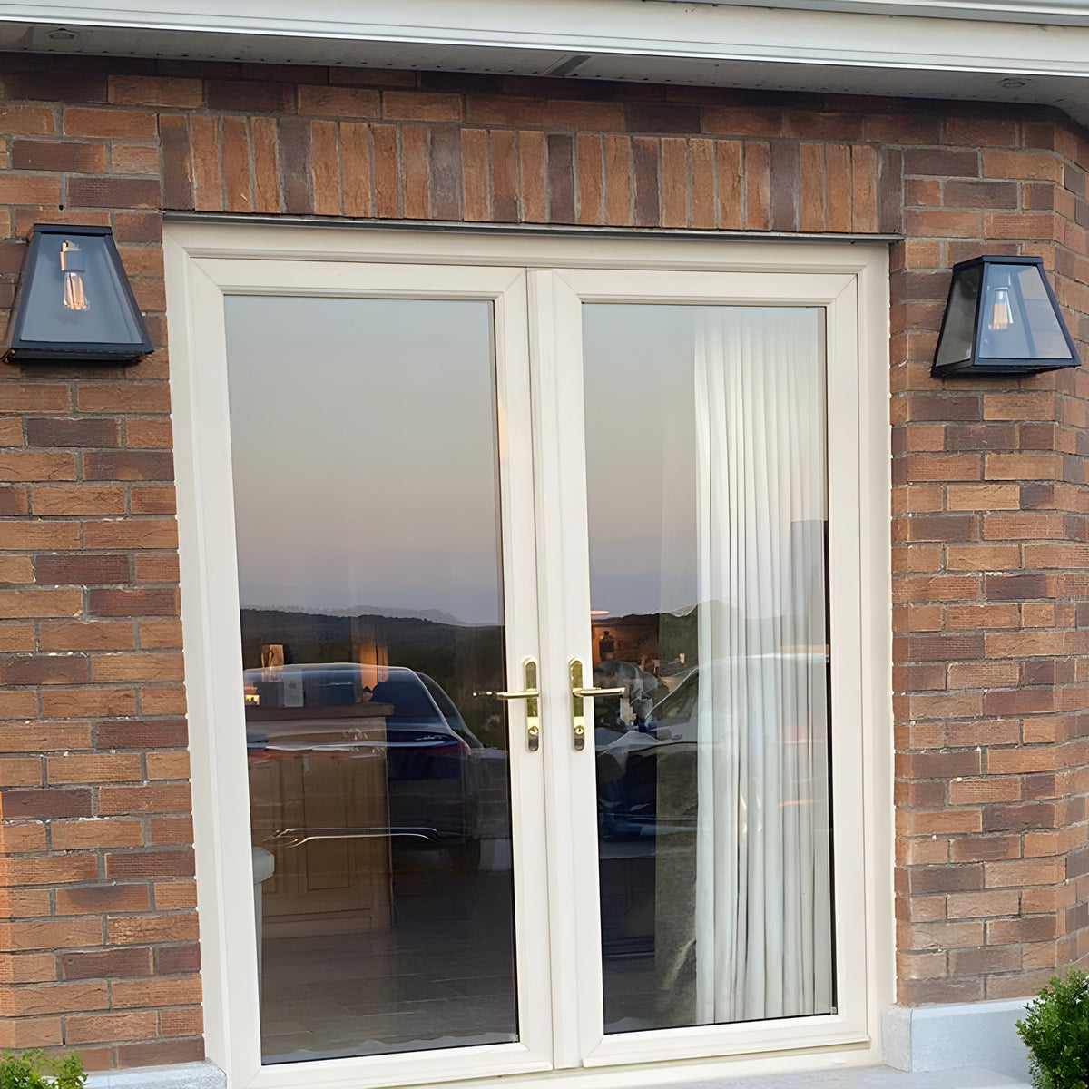 Contemporary galvanised steel bulkhead wall lights in black flank a French door on a brick exterior, each with a tapered glass shade housing an exposed bulb.