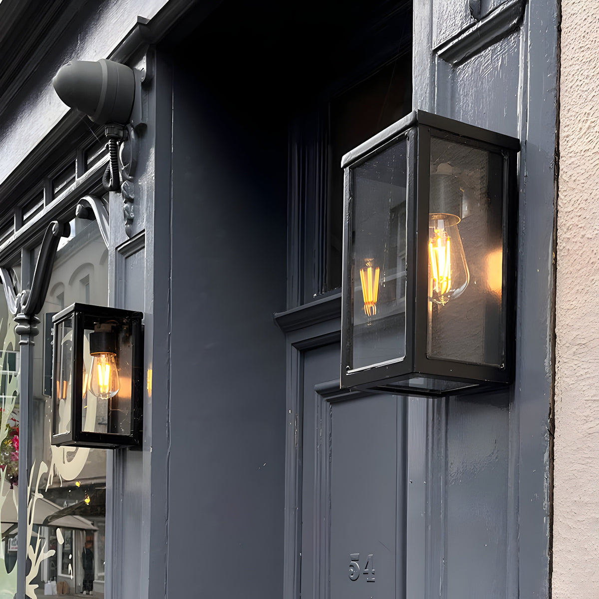 Contemporary Box Wall Lantern in black with clear glazing and exposed bulb, mounted beside a grey doorway on a shopfront exterior.