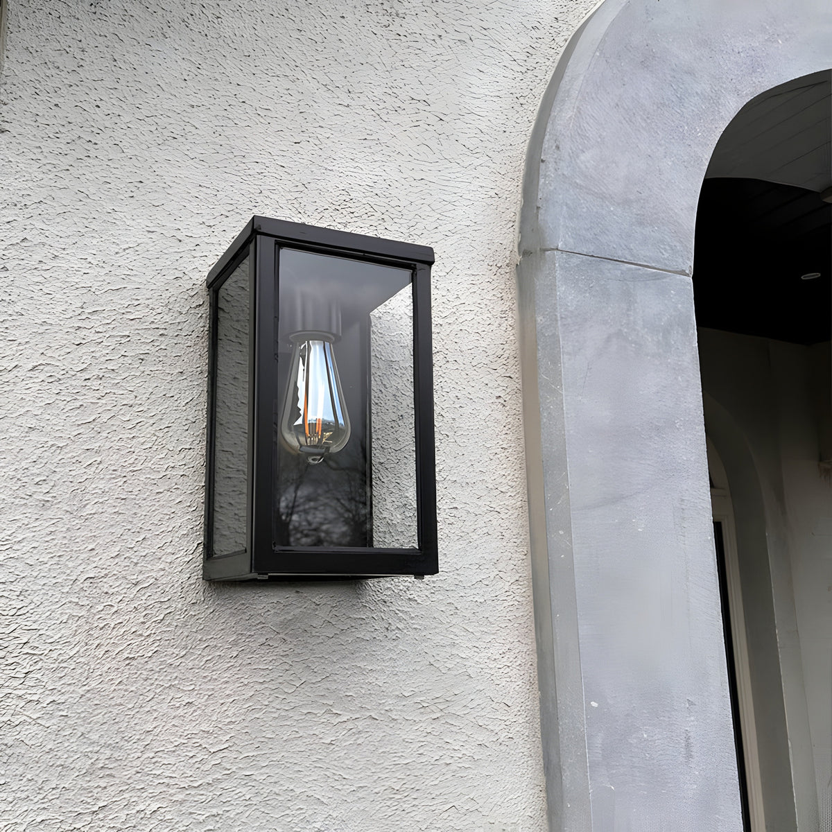 Contemporary Box Wall Lantern in black steel with clear glazing and an exposed E27 bulb, mounted flat on a textured white wall beside an arched stone doorway.