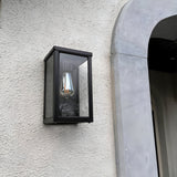 Contemporary Box Wall Lantern in black steel with clear glazing and an exposed E27 bulb, mounted flat on a textured white wall beside an arched stone doorway.