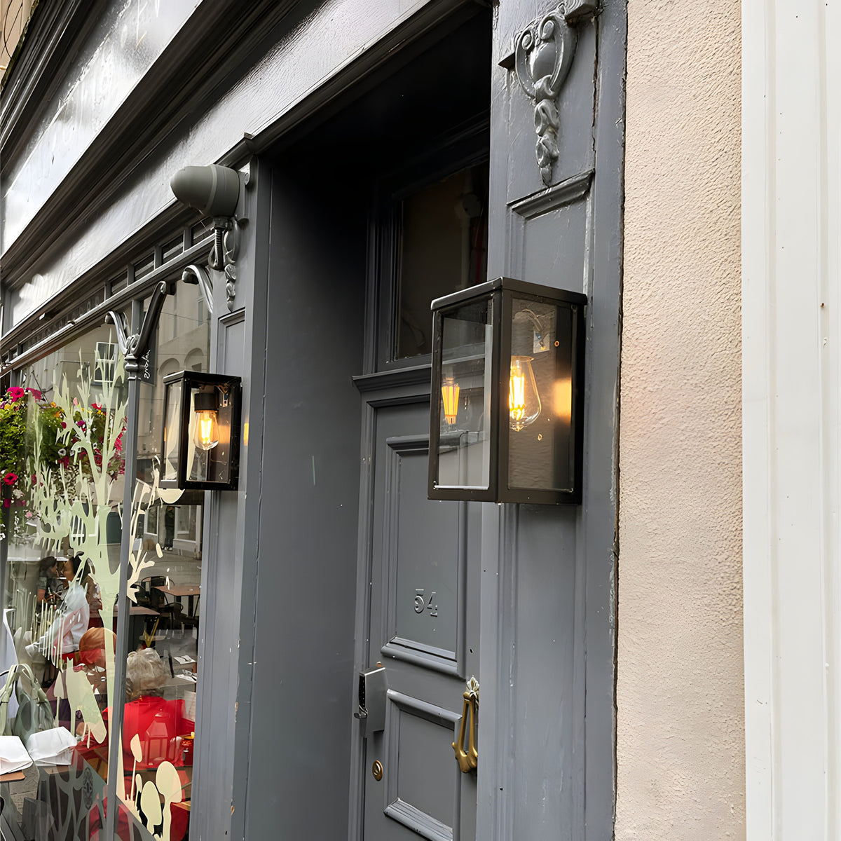 Contemporary Box Wall Lantern in black, a squared glass-panelled outdoor light with visible bulb, mounted beside a grey period door on a shopfront exterior.