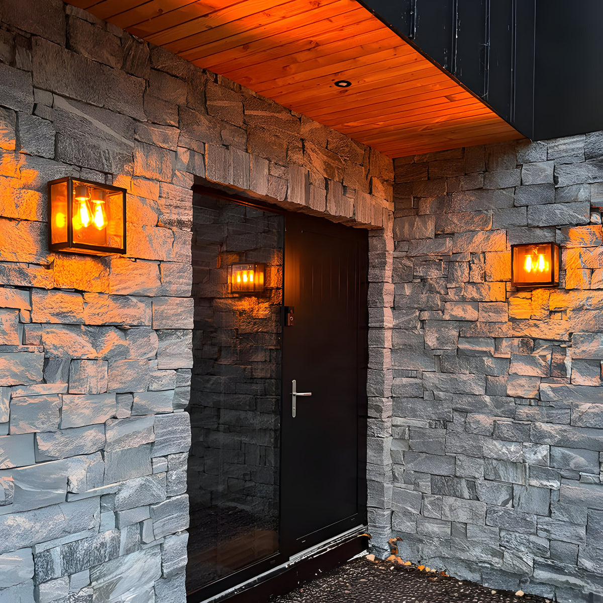 Contemporary Square Box Wall Lantern in black steel with glass panels, mounted on a stone wall beside a dark front door, glowing with warm bulb light.