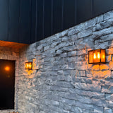Contemporary Square Box Wall Lantern in black steel with glass panel, mounted on a stone wall beside a matching smaller lantern by a dark front door, warm bulb glow visible.