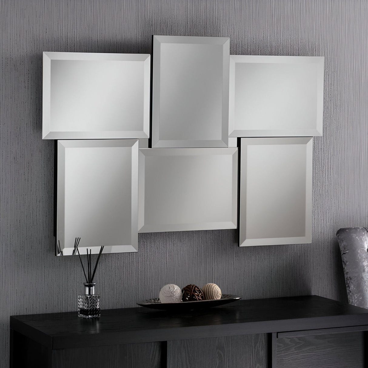 The 112x79cm Verra Rectangle Wall Mirror, made up of six staggered rectangular panels, hangs gracefully on a textured gray wall above a dark wood console table. In this modern bedroom setup, the table is adorned with decorative items such as a reed diffuser and spherical ornaments.