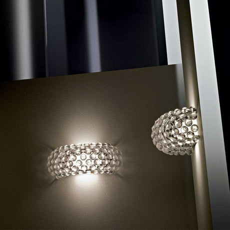 The Briller LED Large Wall Light - Gold, with its cluster of crystal-like beads and luxurious gold finish, reflects off a nearby glass surface to create a soft, ambient glow. Set against a neutral background, this energy-efficient fixture enhances its luminous appearance.