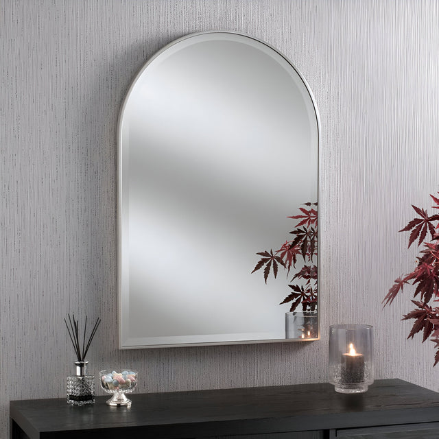 The Bellagio Arched Wall Mirror Silver - 93x63cm hangs on a textured gray wall above a black console table, which holds a glass vase with red leaves, a lit candle, a reed diffuser, and a small jar of candy.