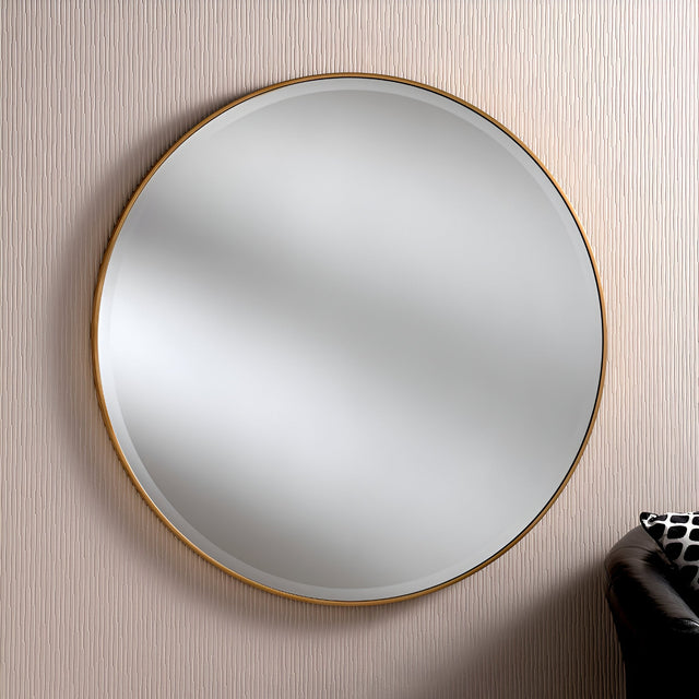 The Camden Circle Wall Mirror in gold, boasting a minimalist design, is mounted on a textured, light-colored wall. In the bottom right corner, a section of a black leather sofa adorned with a patterned cushion can be seen.