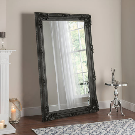 A black Chantelle leaner mirror with baroque elegance is propped against a beige wall near a window with sheer curtains. Beside it, a silver round side table holds a reed diffuser, while on the white mantel, a lit candle provides a gentle glow. The floor is adorned with a patterned blue and white rug.