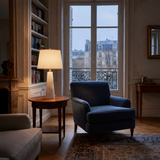 A cozy living room features a blue armchair, the Orix 1 Light Pillar Alabaster Table Lamp – Soft White on a round wooden side table, built-in bookshelves, a fireplace, and a large window overlooking Parisian buildings at dusk.