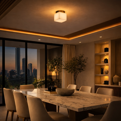 A modern dining room with a marble table, cream chairs, built-in shelves, large windows showing a sunset city skyline, and illuminated by the Bravora 1 Light Cube Alabaster Semi-Flush Ceiling Light in Soft White & Brushed Brass.