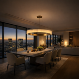 A modern dining room features the Lucenza LED Small Halo Ring Alabaster Chandelier in brushed brass & soft white above a long table with cream chairs, floor-to-ceiling windows overlooking a sunset skyline, and warm lighting from candles and plants.