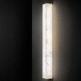 The Valmera LED Large Slim Vertical Alabaster Wall Light in Soft White emits a gentle, diffused glow and looks elegant when mounted on a dark textured wall.