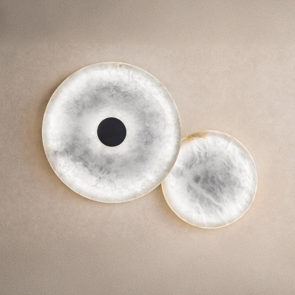 The Solmira LED Dual Disc Alabaster Wall Light in soft white and brushed brass features two overlapping sculptural discs, one with a dark center, mounted on a textured beige wall.