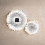 The Solmira LED Dual Disc Alabaster Wall Light in soft white and brushed brass features two overlapping sculptural discs, one with a dark center, mounted on a textured beige wall.
