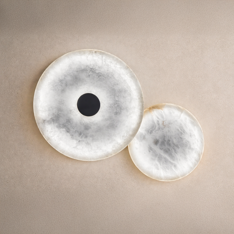 The Solmira LED Dual Disc Alabaster Wall Light in soft white and brushed brass features two overlapping sculptural discs, one with a dark center, mounted on a textured beige wall.
