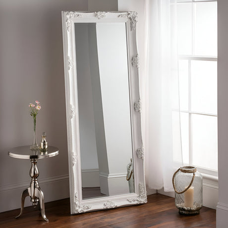 The Luxia Leaner Mirror in white, measuring 171x69cm, rests against a wall near a tall window adorned with sheer curtains. A small round table holding a decorative jar and flowers is situated nearby, alongside a large glass jar on the floor. The room boasts a wooden floor that harmonizes beautifully with the elegant leaner mirror.