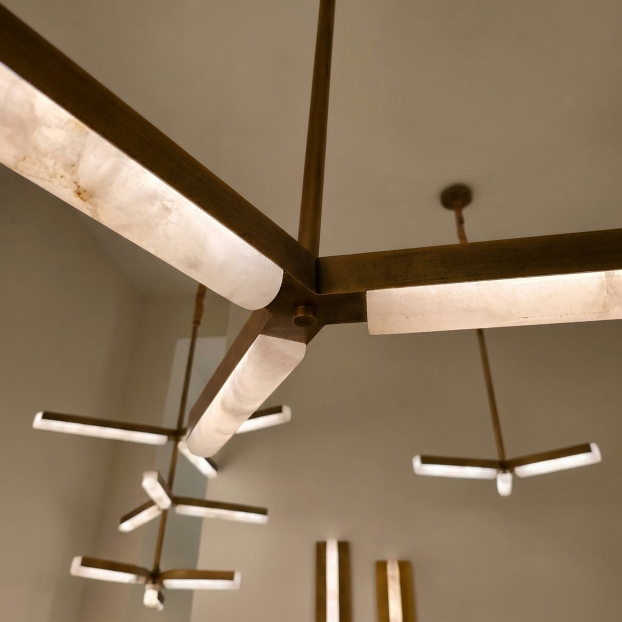 Elvaris LED Radial Alabaster Chandelier in brushed brass, close-up of the central hub with three radiating alabaster arms glowing warm white, additional fixtures visible in a neutral room setting.
