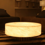 Alurien LED large alabaster coffee table in soft white, round drum shape glowing warmly on a recessed base, shown in a dim living room setting.