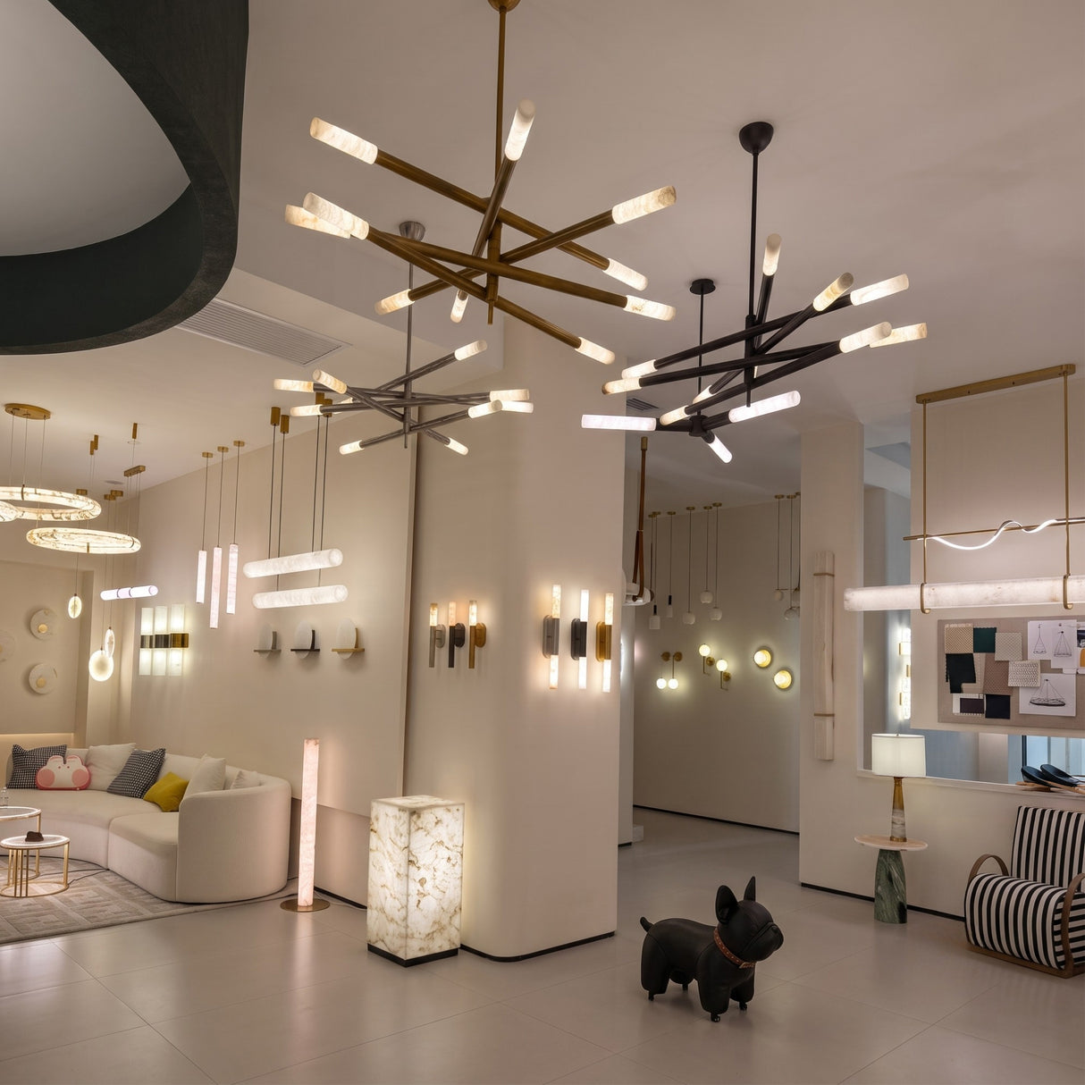 Aurelian LED chandeliers with crossed copper arms in satin nickel finish and cylindrical alabaster diffusers, shown suspended over a showroom seating and display area.