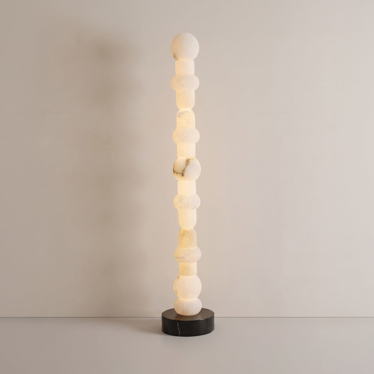 Stratalux LED floor lamp with stacked alabaster spheres and discs glowing warm white, set on a round matt black base against a plain neutral background.