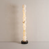 Stratalux LED floor lamp with stacked alabaster spheres and discs glowing warm white, set on a round matt black base against a plain neutral background.