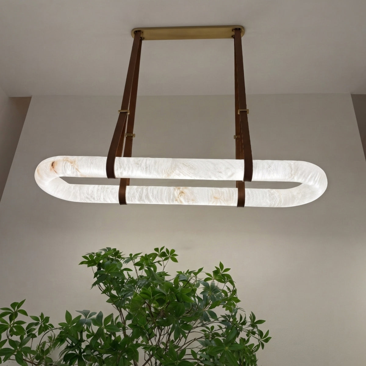 Cintura LED Small Alabaster Floating Pendant Light with oval stone ring, brass ceiling plate and brown leather straps, shown illuminated above a green plant in a neutral room.