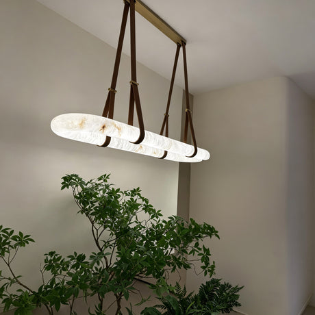 Cintura LED Small Alabaster Floating Pendant Light in soft white, oval alabaster ring suspended by brown leather straps from a copper ceiling bar, shown illuminated above indoor plants.