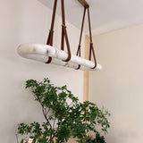 Cintura LED Small Alabaster Floating Pendant Light in soft white, an oval alabaster ring hung by brown leather straps from a copper ceiling bar, shown above greenery in a home setting.