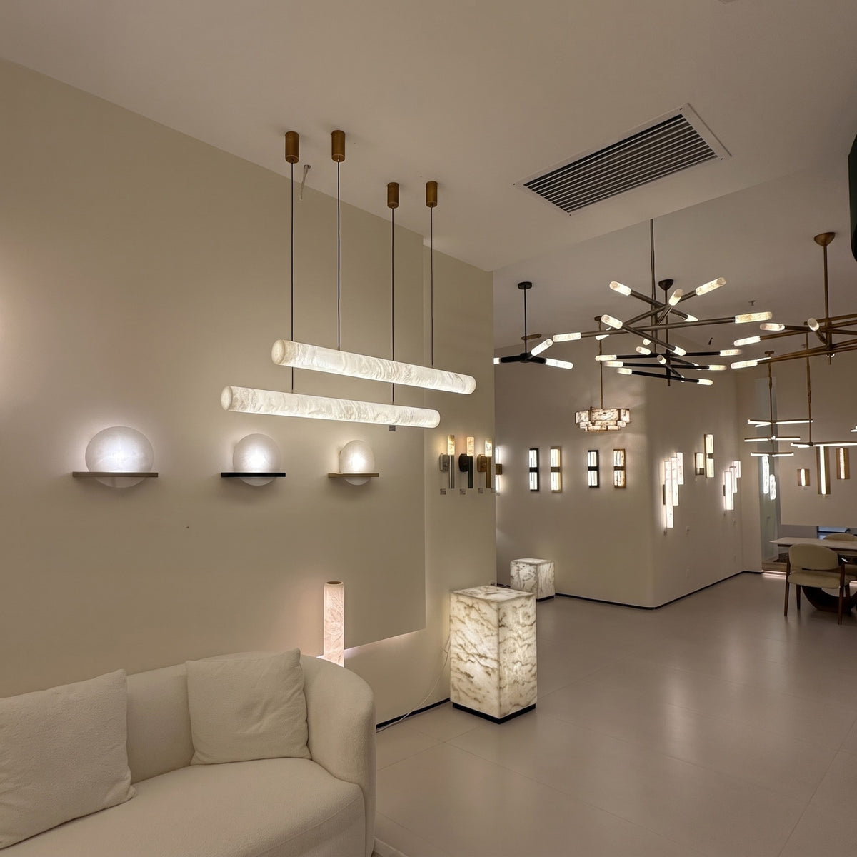 Zareon LED Large Linear Alabaster Chandelier in soft white, shown as two cylindrical pendants on slim cables above a showroom seating area with warm ambient glow.