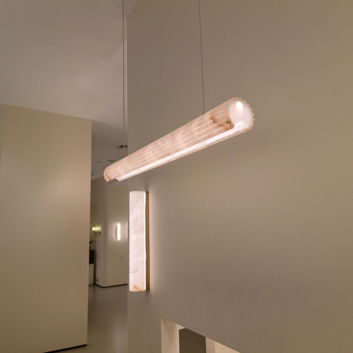 Zenvia LED Alabaster Linear Chandelier in soft white, a cylindrical pendant suspended from a cable, glowing warm white, shown near a matching alabaster wall light in a neutral interior.