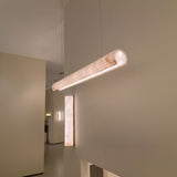 Zenvia LED Alabaster Linear Chandelier in soft white, a cylindrical pendant suspended from a cable, glowing warm white, shown near a matching alabaster wall light in a neutral interior.
