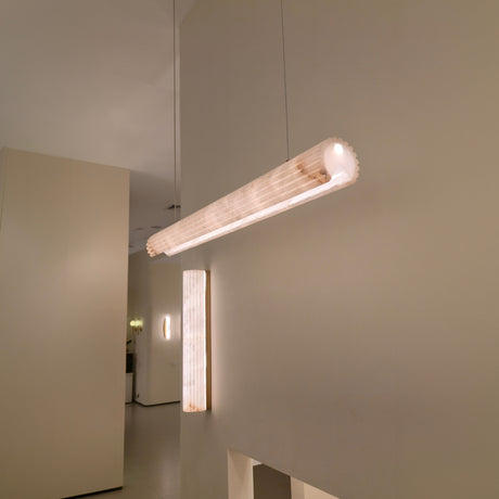 Zenvia LED Alabaster Linear Chandelier in soft white, a cylindrical pendant suspended from a cable, glowing warm white, shown near a matching alabaster wall light in a neutral interior.