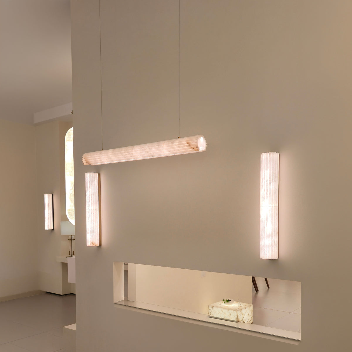 Zenvia LED Alabaster Linear Chandelier in soft white, a horizontal cylindrical pendant suspended from cables, shown alongside matching alabaster wall lights in a hallway setting.