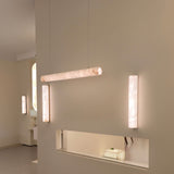 Zenvia LED Alabaster Linear Chandelier in soft white, a horizontal cylindrical pendant suspended from cables, shown alongside matching alabaster wall lights in a hallway setting.