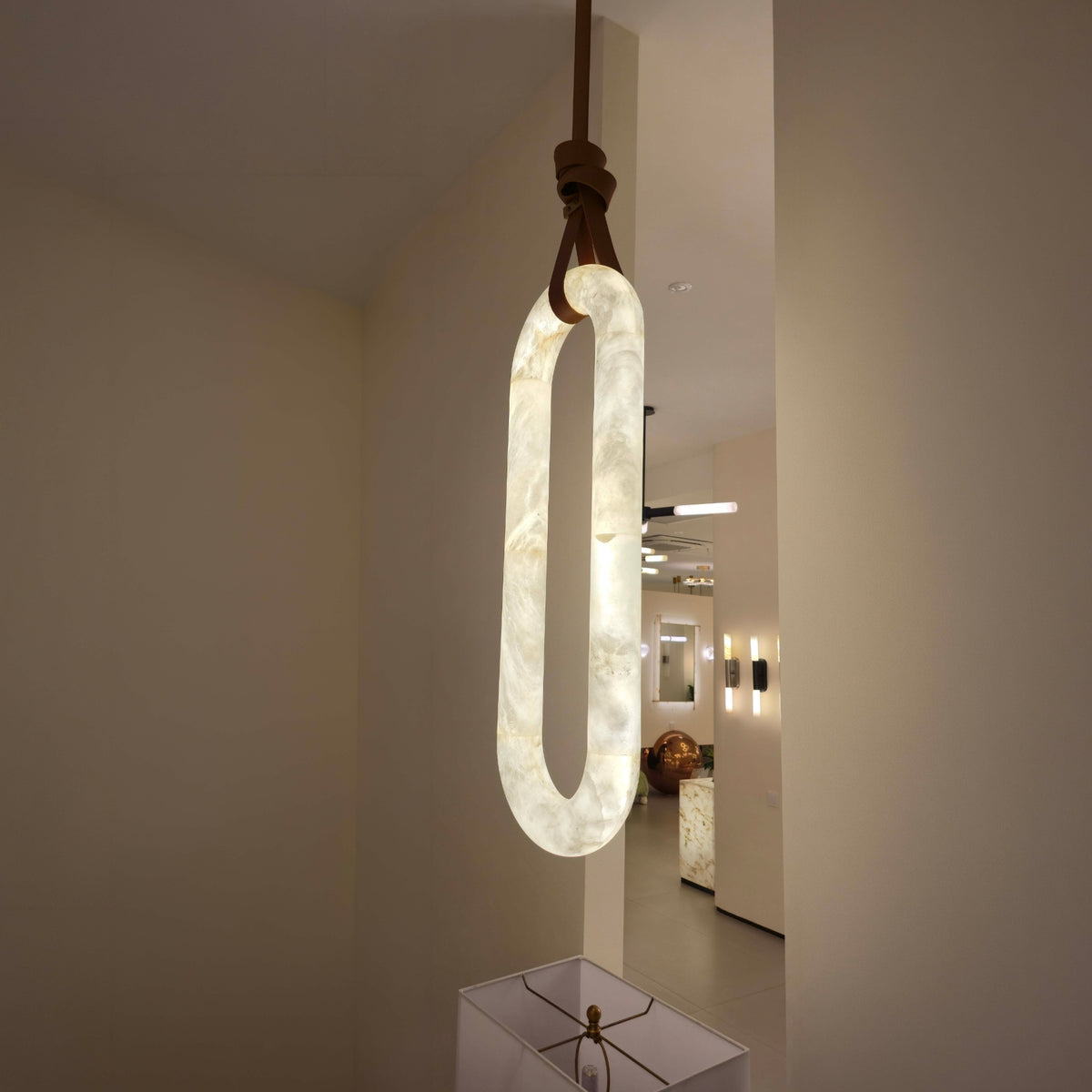 Serava Large LED Alabaster Pendant Light in Soft White, an elongated capsule-shaped fixture with copper leather strap detail, glowing softly in a hallway setting.