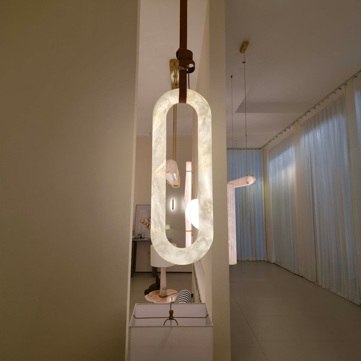 Serava Small alabaster pendant light with elongated rounded form and brown leather strap detail, shown glowing warm white in a hallway setting.