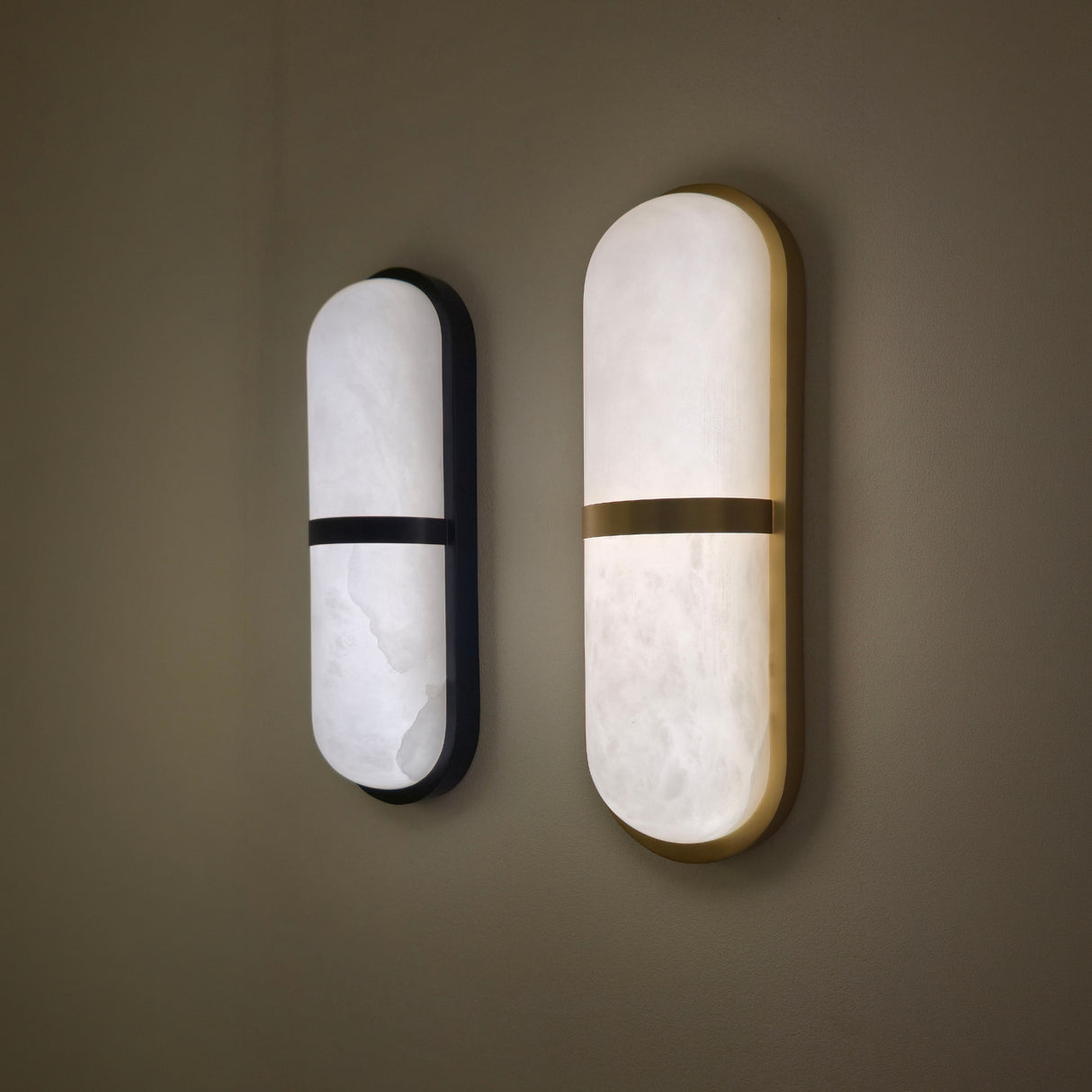 Zantari LED alabaster wall light in brushed brass, capsule-shaped natural stone panel with horizontal brass band, shown beside a matte black version on a wall.
