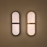 Zantari LED Alabaster Wall Light in brushed brass, capsule-shaped with a horizontal brass band, shown lit beside a matte black version on a plain wall.