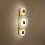 Calyra LED Alabaster Wall Light with three hand-carved white alabaster forms on a brass spine, each with a central brass disc, glowing warm white on a plain wall.