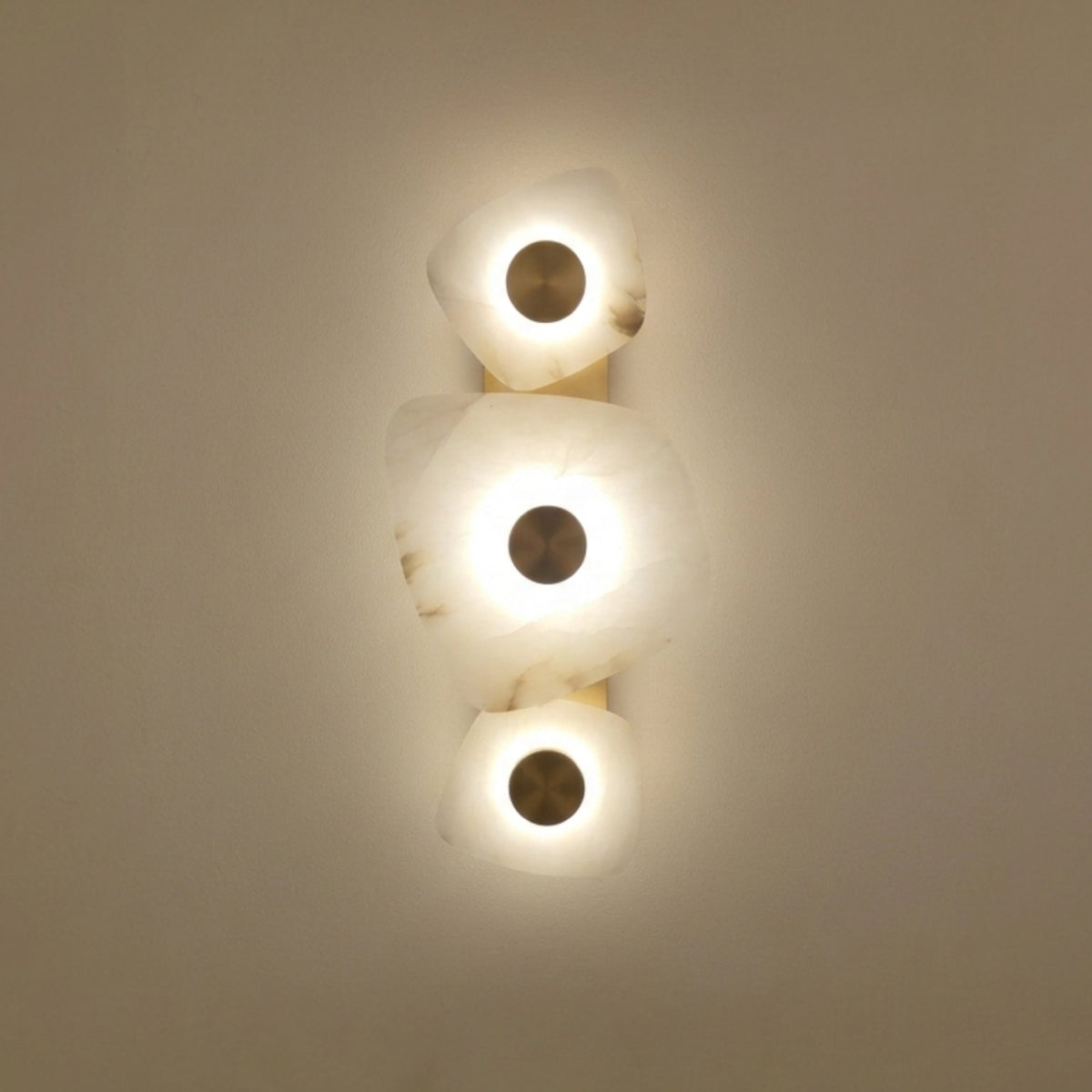 Calyra LED Alabaster Wall Light with three hand-carved white alabaster forms on a slim brass spine, each glowing warmly around a central brass disc, shown lit against a plain background.