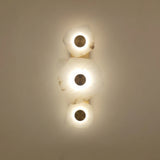Calyra LED Alabaster Wall Light with three hand-carved white alabaster forms on a slim brass spine, each glowing warmly around a central brass disc, shown lit against a plain background.