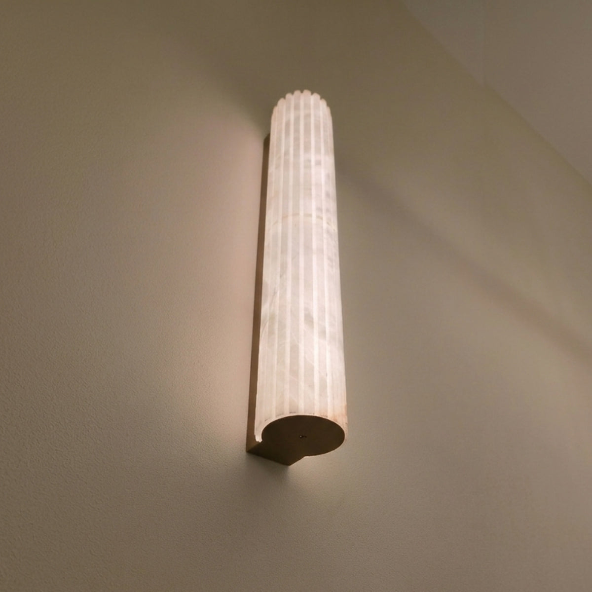 Zenvia LED Alabaster Wall Light with fluted natural stone column and brushed brass back plate, illuminated warm white on a plain wall.