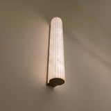 Zenvia LED Alabaster Wall Light with fluted natural stone column and brushed brass back plate, illuminated warm white on a plain wall.