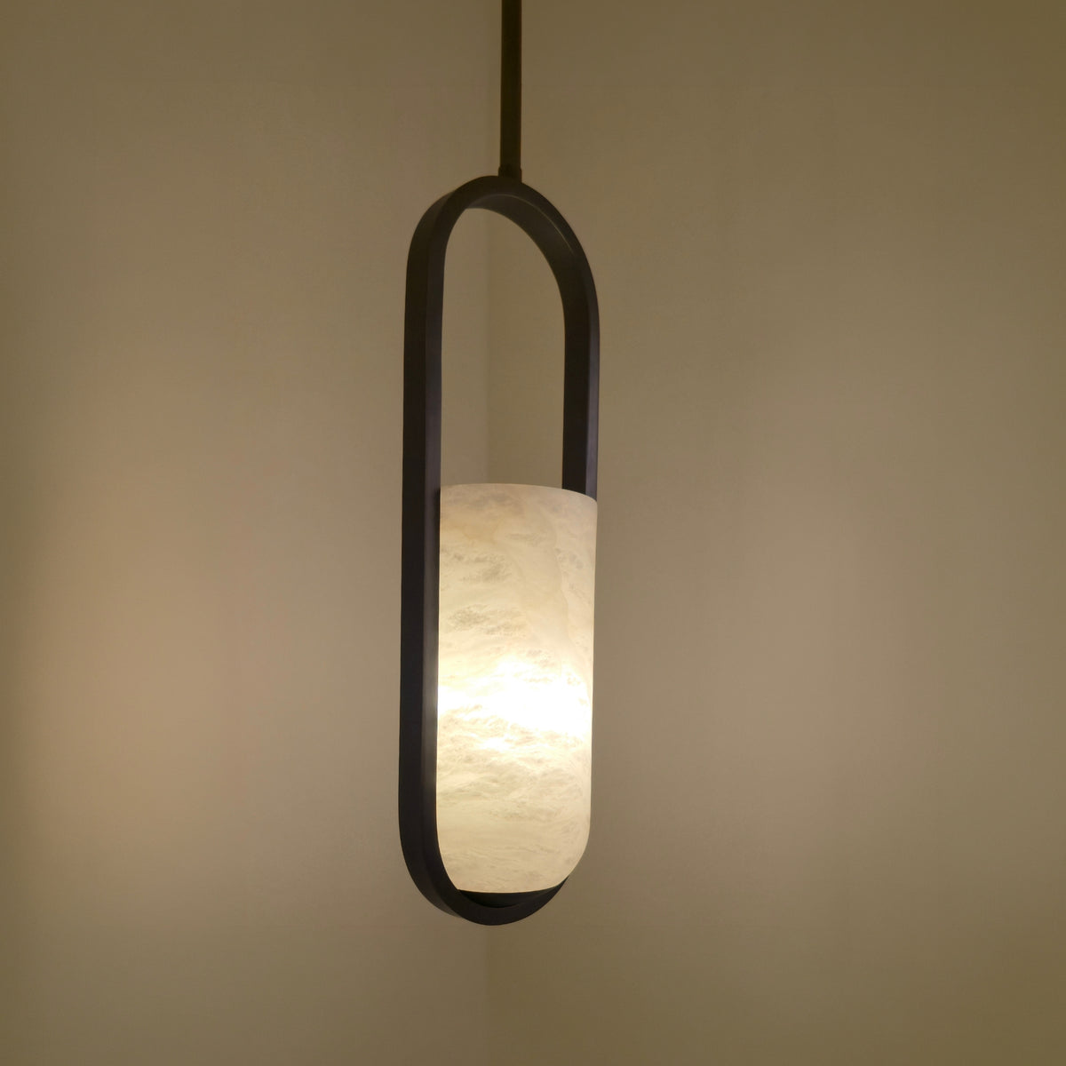 Eliona Capsule Alabaster Pendant Light in matt black, a single cylindrical alabaster shade lit warmly within an oval capsule frame, shown against a neutral wall.