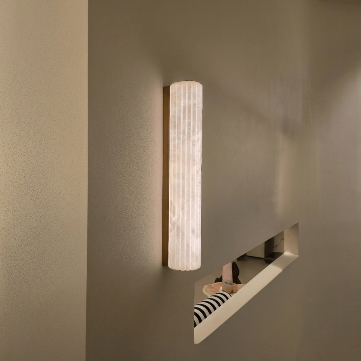 Zenvia LED Alabaster Wall Light in Brushed Brass, a slim vertical fixture with fluted natural alabaster column, shown illuminated on a grey wall in a hallway setting.
