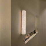 Zenvia LED Alabaster Wall Light in Brushed Brass, a slim vertical fixture with fluted natural alabaster column, shown illuminated on a grey wall in a hallway setting.