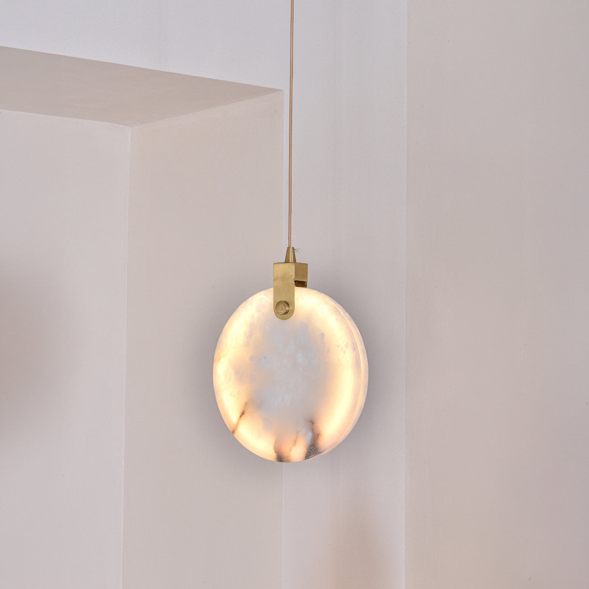Luna LED Alabaster Disc Pendant Light 200mm in brushed brass, glowing warm white, hung by a brass hinge and cable in a light-toned room corner.